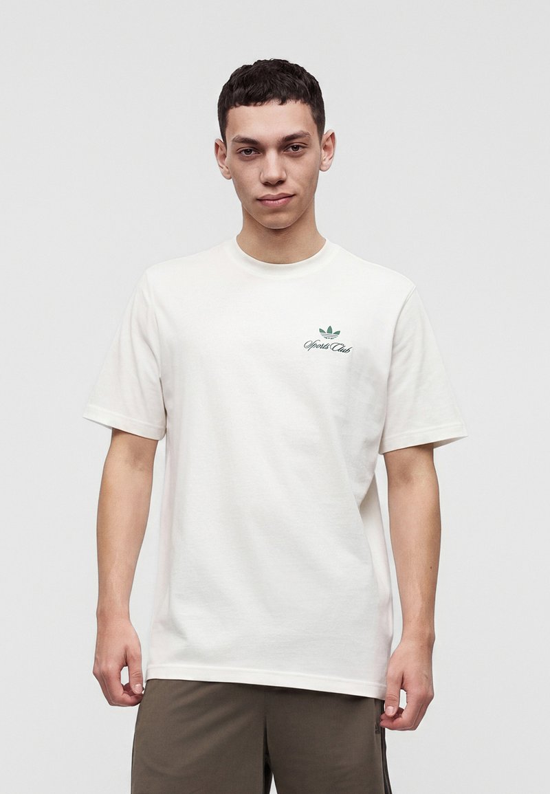 Junger Mann trägt ein weißes kurzärmeliges T-Shirt mit kleinem grünem Sportlogo auf der linken Brust und dunkle Hose, steht vor einem einfarbigen Hintergrund.