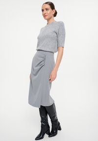 Grauer gestrickter Pullover mit kurzen Ärmeln und Knopfverzierungen, kombiniert mit einem grauen gerafften Midikleid und kniehohen schwarzen Lederstiefeln.