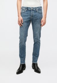 Ανοιχτό μπλε τζιν με εφαρμοστή σχεδίαση, κατασκευασμένο από ύφασμα τζην. Διαθέτει κλείσιμο με κουμπί και πέντε τσέπες. Φοριέται με μαύρες καουμπόικες μπότες.