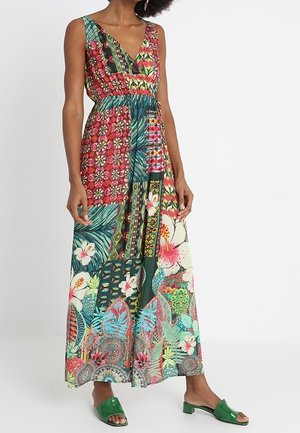 Femme portant une robe longue patchwork sans manches avec un col en V et des motifs floraux et géométriques, associée à des sandales vertes à talons bas et à bouts ouverts.