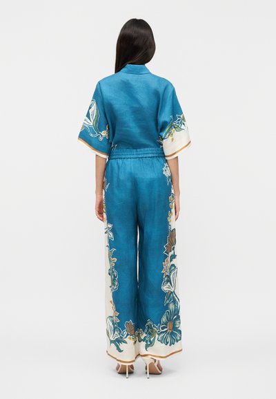 Γυναίκα με μακριά σκούρα μαλλιά, φορώντας ένα ολόσωμο παντελόνι σε στυλ παντελονιού με φαρδιά πόδια, με τυπωμένο ανθικό σχέδιο σε πετρόλ χρώμα, στέκεται με την πλάτη προς την κάμερα σε απλό φόντο.