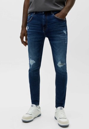 Homme portant un jean slim bleu déchiré aux genoux et des baskets blanches, debout avec une main dans la poche.
