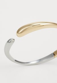Bracelete de pulso em metal com um design curvado, apresentando um acabamento brilhante em ouro e prata, textura suave e detalhes de acentuação sutis.