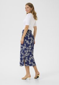 Jupe midi floral bleu marine avec une fente haute, taille élastique et motif floral blanc. Associée à un haut blanc ajusté et des sandales à talons jaunes.