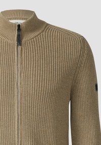 Beige gestrickter Reißverschluss-Pullover mit hohem Kragen, der ein strukturiertes Rippdesign und einen schwarzen Logo-Akzent am Ärmel aufweist.