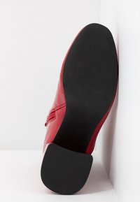 Bottine rouge à cheville avec semelle en caoutchouc noire et petit talon, positionnée semelle vers le haut contre un fond d'angle blanc.