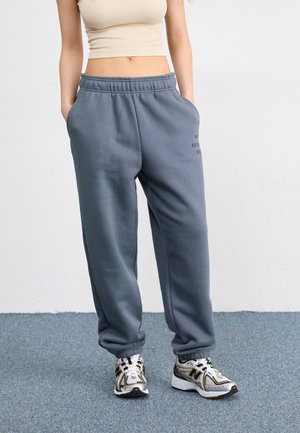 Pantalones de chándal grises de corte relajado, con cinturilla elástica, bolsillos laterales y detalle de logo impreso. Llevados con zapatillas de correr con acentos dorados.