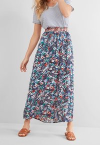 Femme portant une jupe maxi bleue à fleurs avec une taille froncée, associée à un t-shirt à rayures blanches et bleues marines à manches courtes et des sandales marron.
