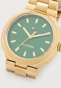 Złoty zegarek na nadgarstek z zieloną tarczą, złotymi znacznikami godzin i wskazówkami, logo Tommy Hilfiger oraz ogniwowym metalowym paskiem.