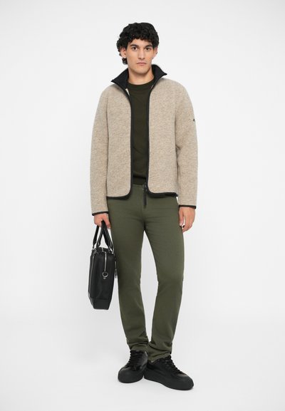 Beige zip-up jacket with black trim, green shirt underneath, olive green pants, and black shoes, holding a black bag. Smooth textures throughout.