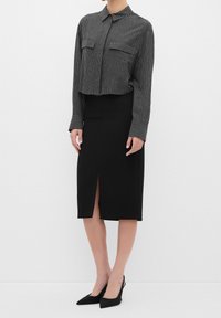 Femme portant un chemisier court gris à fines rayures avec des manches longues et une jupe noire longueur genou avec une fente à l'avant et des talons pointus noirs.