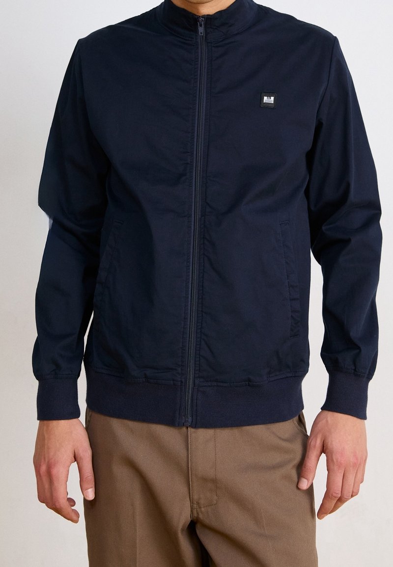 Granatowa kurtka zapinana na zamek błyskawiczny z wysokim kołnierzem, lekkiego materiału, z elastycznymi mankietami, bocznymi kieszeniami i małą naszywką z logo na piersi.