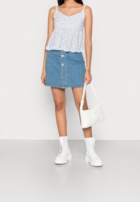 Kvinna iförd en vit blommig ärmlös topp, knäppt denimkjol, vita lace-up stövlar och håller en vit handväska framför en enfärgad bakgrund.