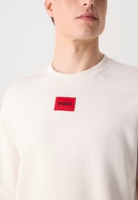 Kreemist sweatshirt, ribaga kaeluses ja punase ristkülikukujulise plaastriga, millel on must tekst "HUGO". Pehme kangas, mõnus lõige.