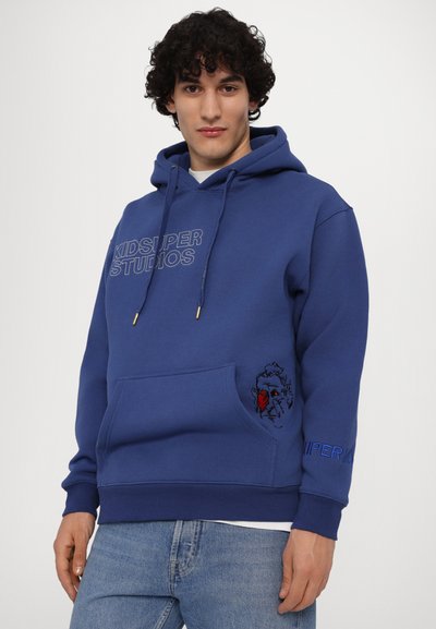Felpa blu con tasca frontale, logo "KIDSUPER STUDIOS", grafica ricamata sulla tasca e lacci con dettagli dorati.