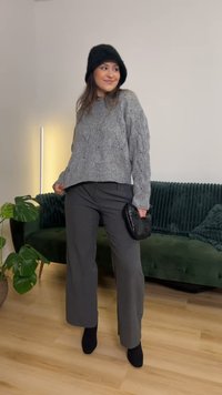 Camisola cinzenta de malha com padrão trançado, calças largas cinzentas, mala preta de mão e botas pretas até ao tornozelo. Planta e sofá verde ao fundo.