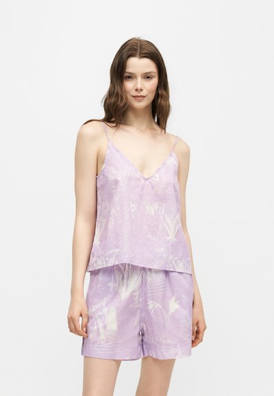 Jeune femme portant un caraco floral violet clair et un short assorti, se tenant devant un fond blanc uni.