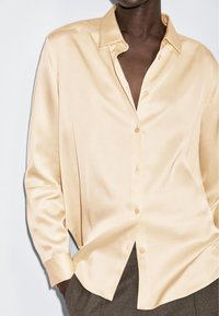Chemise en satin à manches longues couleur beige doux, dotée d'un col pointu, d'une fermeture à boutons sur le devant et d'une coupe ample avec un ourlet légèrement incurvé.