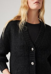 Μαύρο πλεκτό κασκόλ με ριγωτή υφή, v-neck και διακοσμητικά κουμπιά σε σχήμα ασημένιου λουλουδιού. Μακριά μανίκια και μαλακό, παχύ ύφασμα.