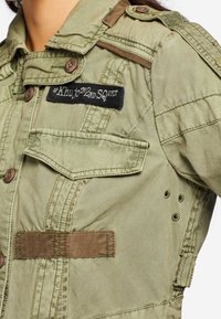 Olivgrön militärinspirerad jacka i lätt tyg, med en svart patch med vit text, flera fickor och bruna detaljer.