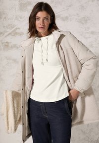 Licht beige gewatteerde jas met een gewatteerde crème binnenvoering, oversized pasvorm. Witte shirt met trekkoorden, donkere jeans en een crème stof in de hand.