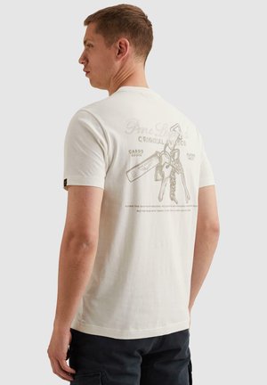 Muž nosiaci biele tričko s vyblednutými potlačenými kľúčmi a textom na tému letectva na chrbte, v kombinácii s čiernymi cargo nohavicami.