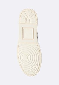 Sole di una scarpa di colore chiaro con modelli di grip testurizzati, tra cui cerchi concentrici, un quadrato e linee in rilievo su uno sfondo semplice.
