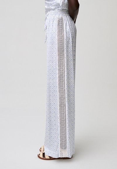 Persona che indossa pantaloni bianchi a gamba larga con stampa floreale blu e dettaglio verticale in pizzo, abbinati a sandali beige con punta aperta, in piedi di lato.