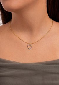 Collar de cadena de oro con un colgante circular adornado con piedras preciosas de colores variados, incluyendo púrpura, rosa y azul, sobre un fondo neutro.