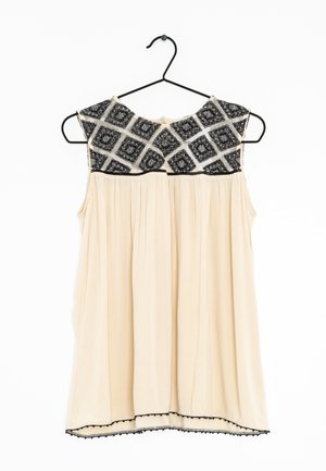 Mouwloze beige blouse met zwart patroon op de schouderpartij, geplisseerd ontwerp en zwarte afwerking langs de zoom. Hangt aan een zwarte haak.