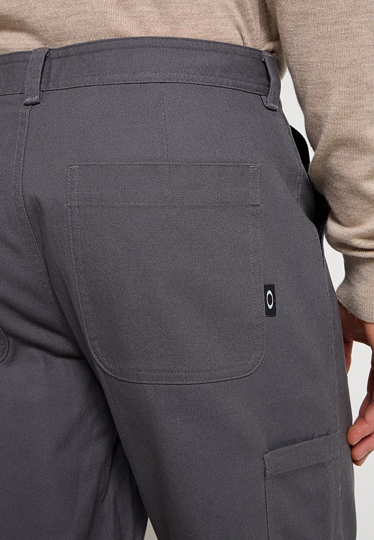 Pantalones cargo grises fabricados en un material duradero que cuentan con un bolsillo trasero y un parche de marca con un logotipo circular. El diseño es práctico y funcional.