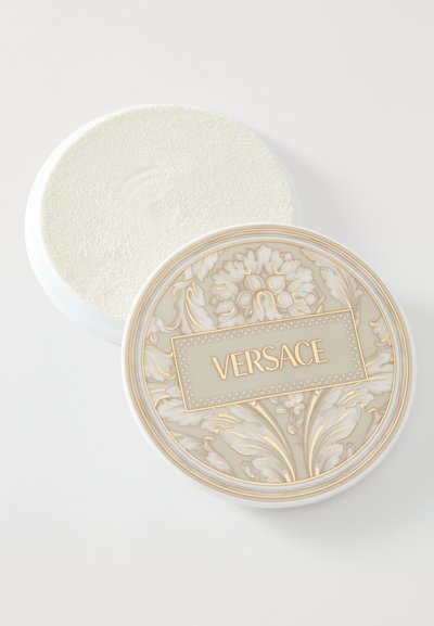Bílá kulatá nádobka s texturovaným krémovým víčkem zdobeným detailními květinovými vzory a zlatým logem Versace uprostřed.