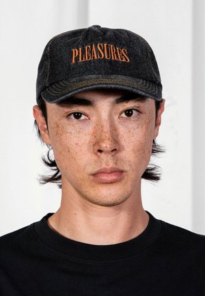 Młoda osoba o jasnej karnacji i piegach, ubrana w czarną czapkę z pomarańczowym napisem "PLEASURES" oraz czarną koszulę, zwrócona przodem na tle białego tła.