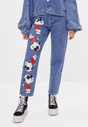 Dżinsy z denimu z powtarzającym się nadrukiem Snoopy’ego w czerwonej koszulce „Joe Cool” wzdłuż lewej nogawki, zestawione z czarnymi trampkami na platformie i kurtką jeansową.