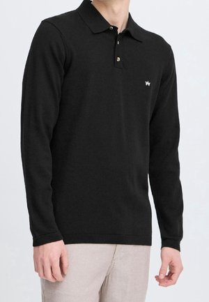 Homme portant un pull polo noir à manches longues avec un petit logo blanc sur la poitrine et un pantalon beige sur fond uni.