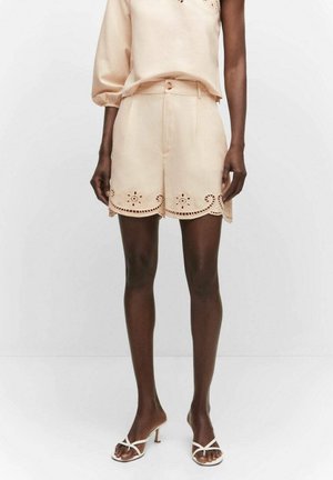 Modelo que lleva pantalones cortos de talle alto beige con dobladillo festoneado de ojales, combinados con un top de una manga a juego y sandalias blancas de tacón con tiras.