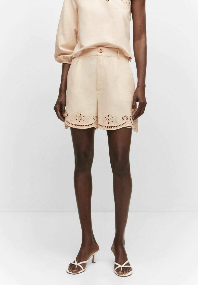 Mannequin portant un short beige taille haute avec un ourlet festonné ajouré, associé à un top asymétrique à manches longues assorti et des sandales blanches à talons avec brides.