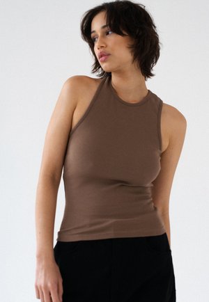 Kobieta o krótkich ciemnych włosach, ubrana w brązowy bezrękawnik i czarne spodnie, patrząca w lewo na białym tle.