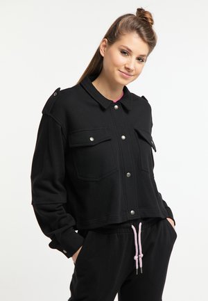Junge Frau mit hellbraunen Haaren im halben Dutt, trägt eine schwarze Jacke mit Knöpfen und schwarze Hosen mit pinken Kordeln, Hände in den Taschen, lächelnd.