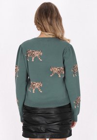 Pulover verde tricotat, cu un model de tigri maro și negri, cu manșete ajustate și tiv zimțat. Este asortat cu o fustă neagră cu volane.
