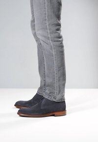 Laivastonsiniset teksturoidut nilkkurit, joissa on sileät ruskeat pohjat, yhdistettynä harmaisiin istuviin farkkuihin. Nilkkureissa on hienovarainen kuvio ja nauhat.