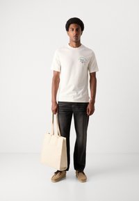 Krämfärgad bomulls-t-shirt med korta ärmar och tryckt grön och blå grafik, kombinerad med mörka jeans och en beige canvas tote-väska.