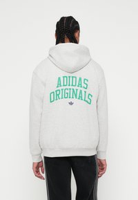 Szara bluza z kapturem z napisem "ADIDAS ORIGINALS" w zielonym kolorze na plecach, z logo poniżej. Teksturowany materiał o luźnym kroju.