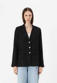 Blazer noir avec texture en jacquard floral, devant à un bouton, quatre boutons décoratifs et deux poches avant. Associé à un jean bleu clair.