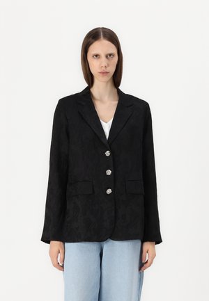 Blazer noir avec texture en jacquard floral, devant à un bouton, quatre boutons décoratifs et deux poches avant. Associé à un jean bleu clair.