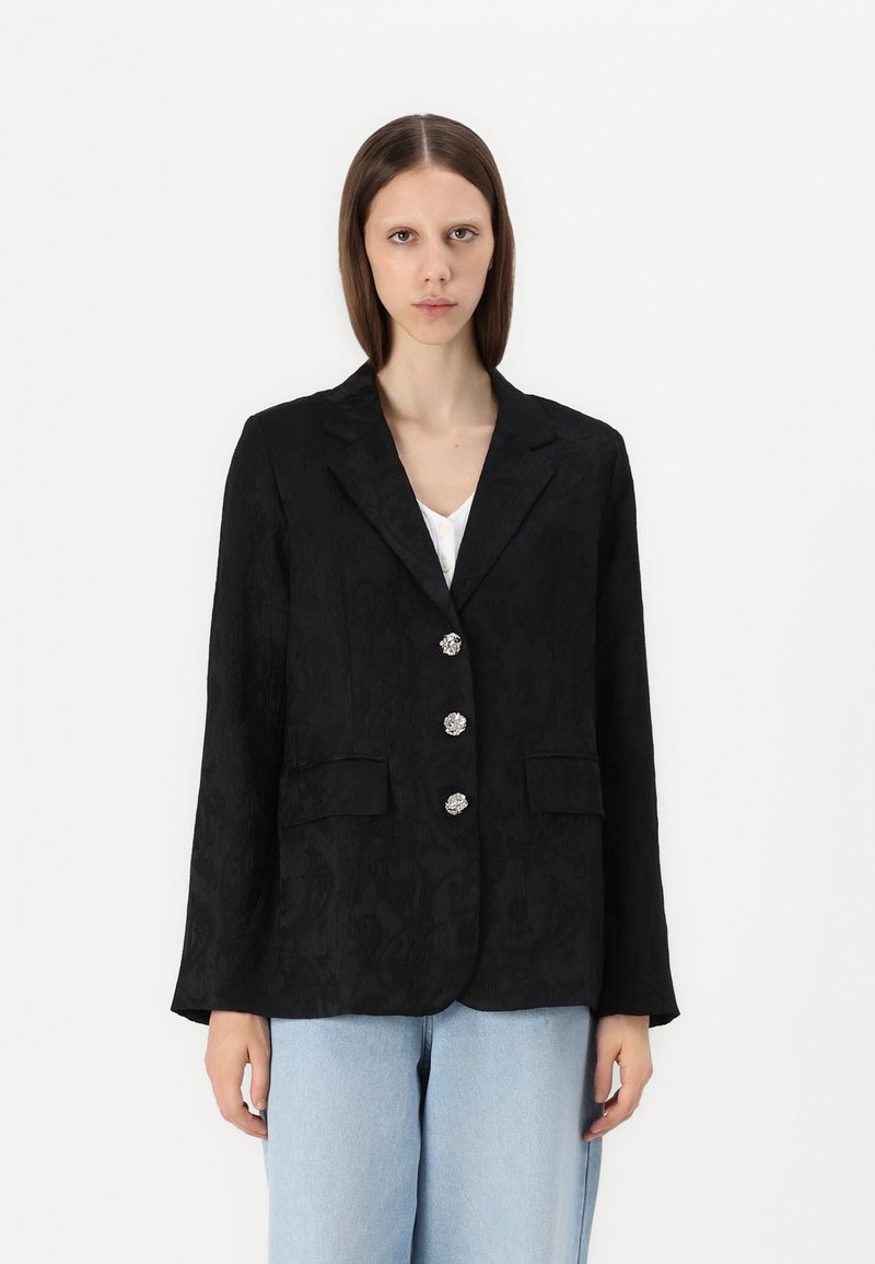 Blazer noir avec texture en jacquard floral, devant à un bouton, quatre boutons décoratifs et deux poches avant. Associé à un jean bleu clair.