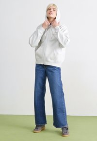 Sweat à capuche zippé gris clair avec une capuche et des poches avant, associé à un jean en denim bleu à jambes larges et des baskets grises sur un sol vert.