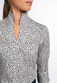 Femme portant un chemisier blanc avec un motif de points noirs irréguliers et un col montant, boutons fermés, manches retroussées, sur fond blanc.