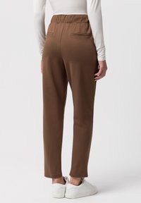 Hnědé tapered kalhoty s elastickým pasem, se dvěma zadními kapsami, v kombinaci s bílými teniskami. Hladká látka, ležérní design.
