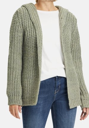 Cardigan en tricot vert avec capuche, décolleté ouvert, motif de point texturé, poignets côtelés, porté sur un haut blanc et un jean en denim.